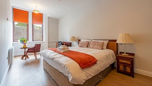 A bedroom with a large bed, two bedside tables, a chair, a window with blinds, and a small table with a vase of flowers. - The Hazlitt Apartments (Paisley)