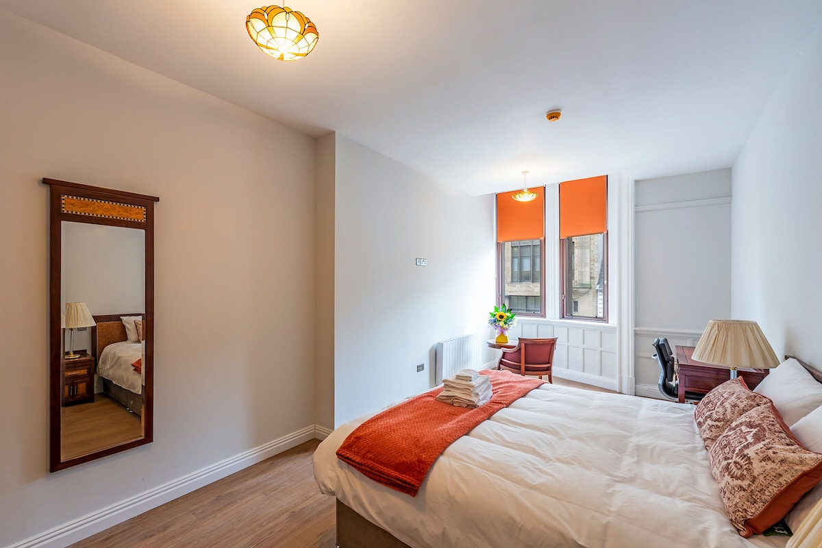 A hotel room with a large bed, a mirror, a window with orange blinds, a desk with a lamp, and a chair.