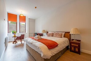 A bedroom with a large bed, two bedside tables, a chair, a window with blinds, and a small table with a vase. - The Hazlitt Apartments (Paisley)