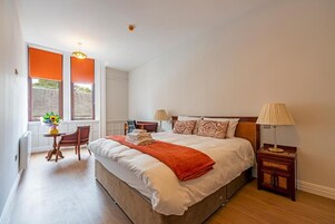 Un dormitorio con una cama grande, dos mesitas de noche, una silla, un ventanal con persianas y una pequeña mesa con un florero.