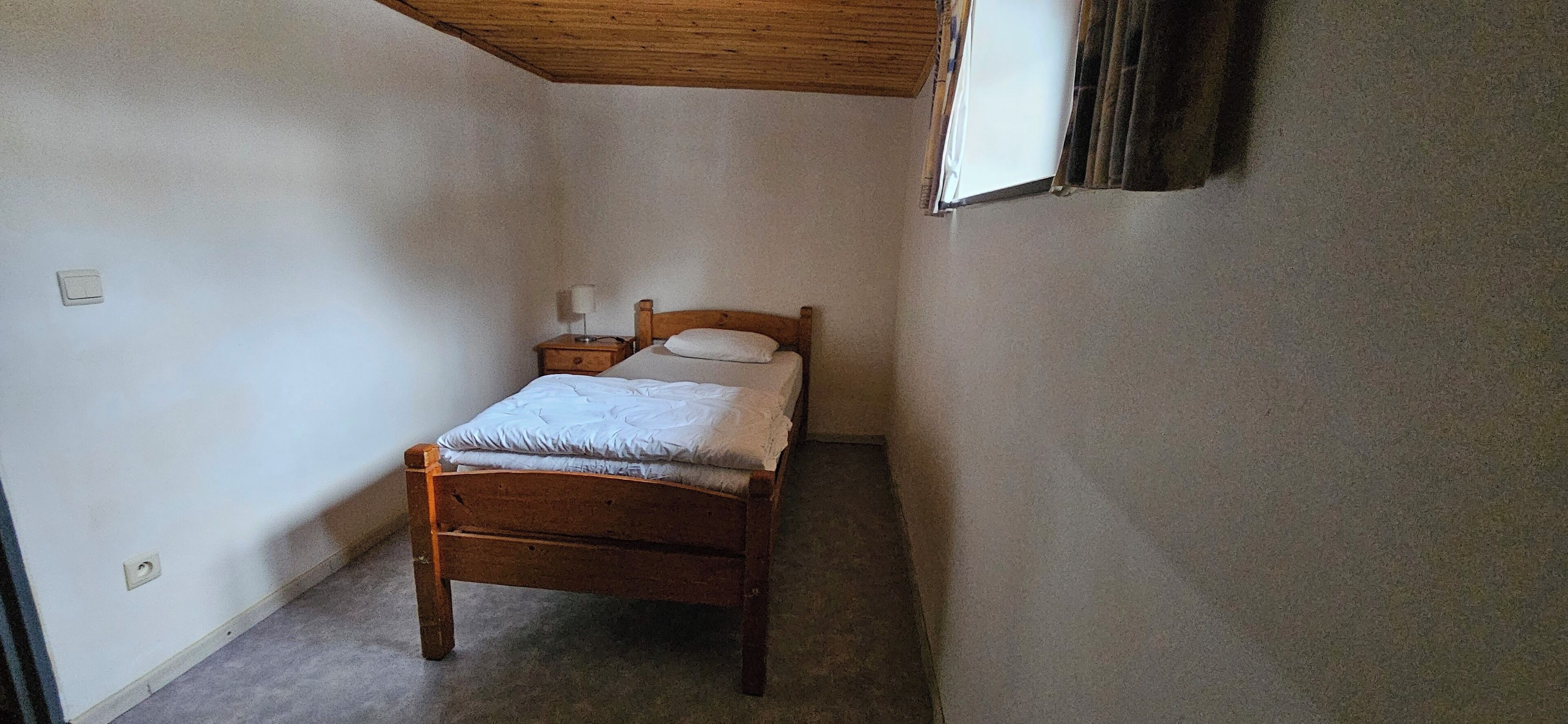 Quarto pequeno com uma cama de solteiro, cabeceira de madeira, mesa de cabeceira com abajur e janela com cortinas.