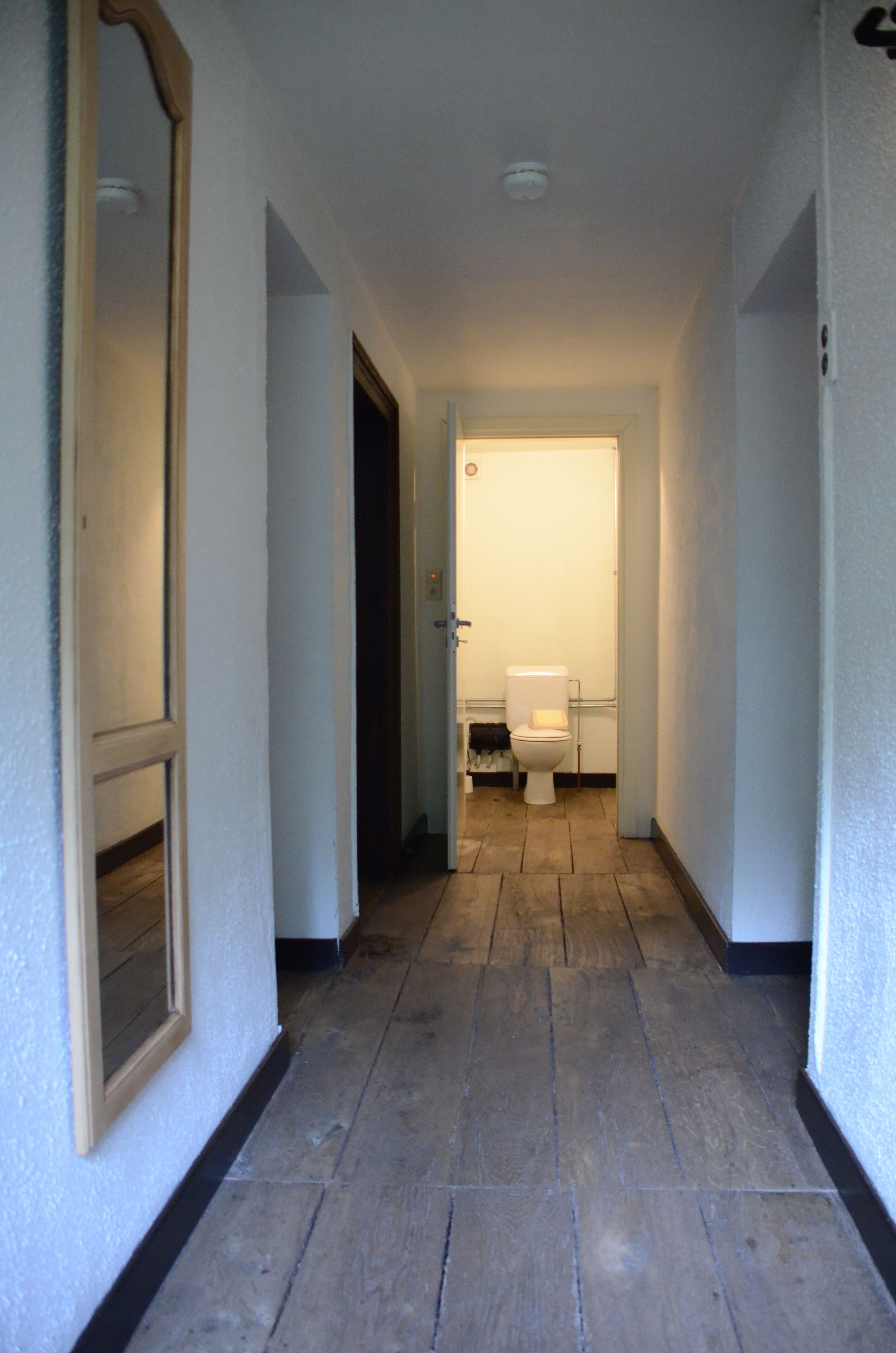 A narrow hallway with a wooden floor, white walls, and a door leading to a bathroom with a toilet and a sink.