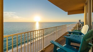 A balcony with a view of the ocean at sunset, featuring a railing and two green Adirondack chairs. - Beachfront 4BR | Heated Pool + Hot Tub (Panama City Beach)