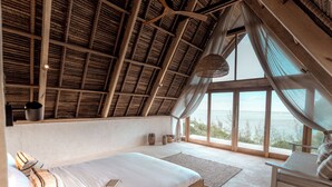 Ein Schlafzimmer mit schrägem Dach, einem großen Fenster, einem Bett, einem Sessel und einer hängenden Deckenleuchte.