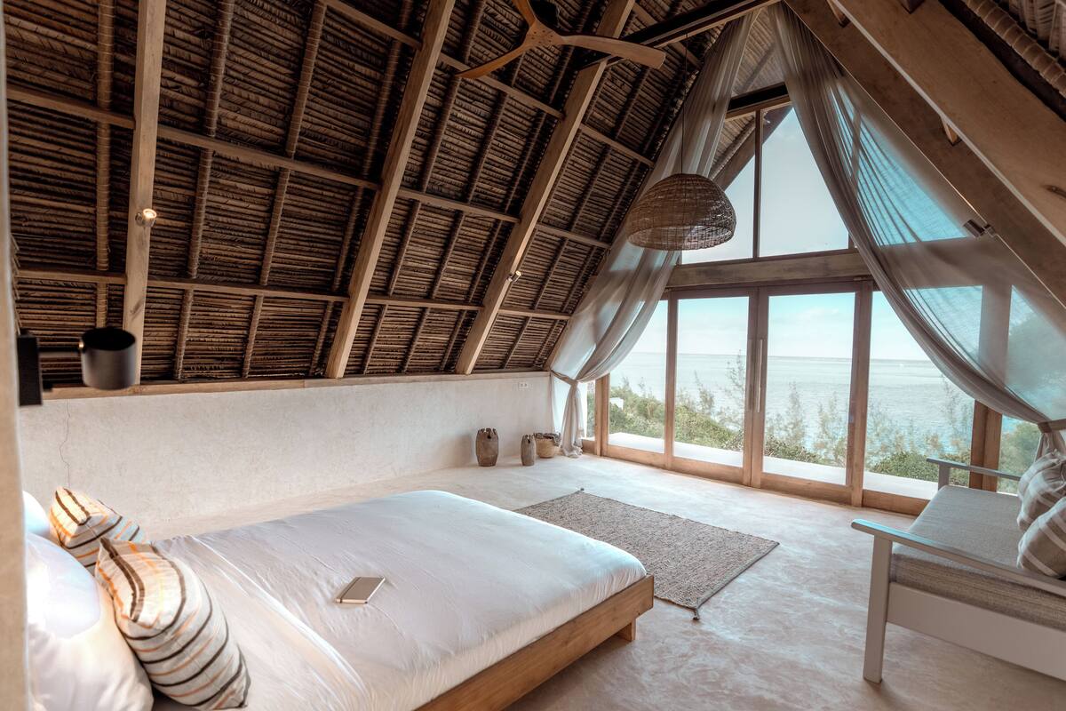 Une chambre avec un plafond en pente, une grande fenêtre, un lit, une chaise et un luminaire suspendu.