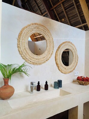 A bathroom with two woven mirrors, a terracotta vase with a plant, a sink with toiletries, and a woven basket with towels. - Tao Resort Madagascar (Antsiranana)