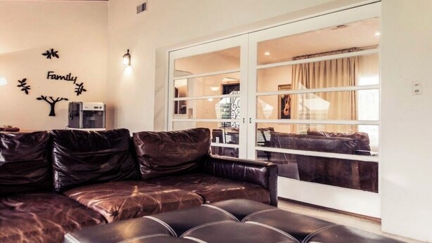 Un salon moderne avec un canapé en cuir marron, un pouf noir et une porte en verre donnant sur un coin repas.