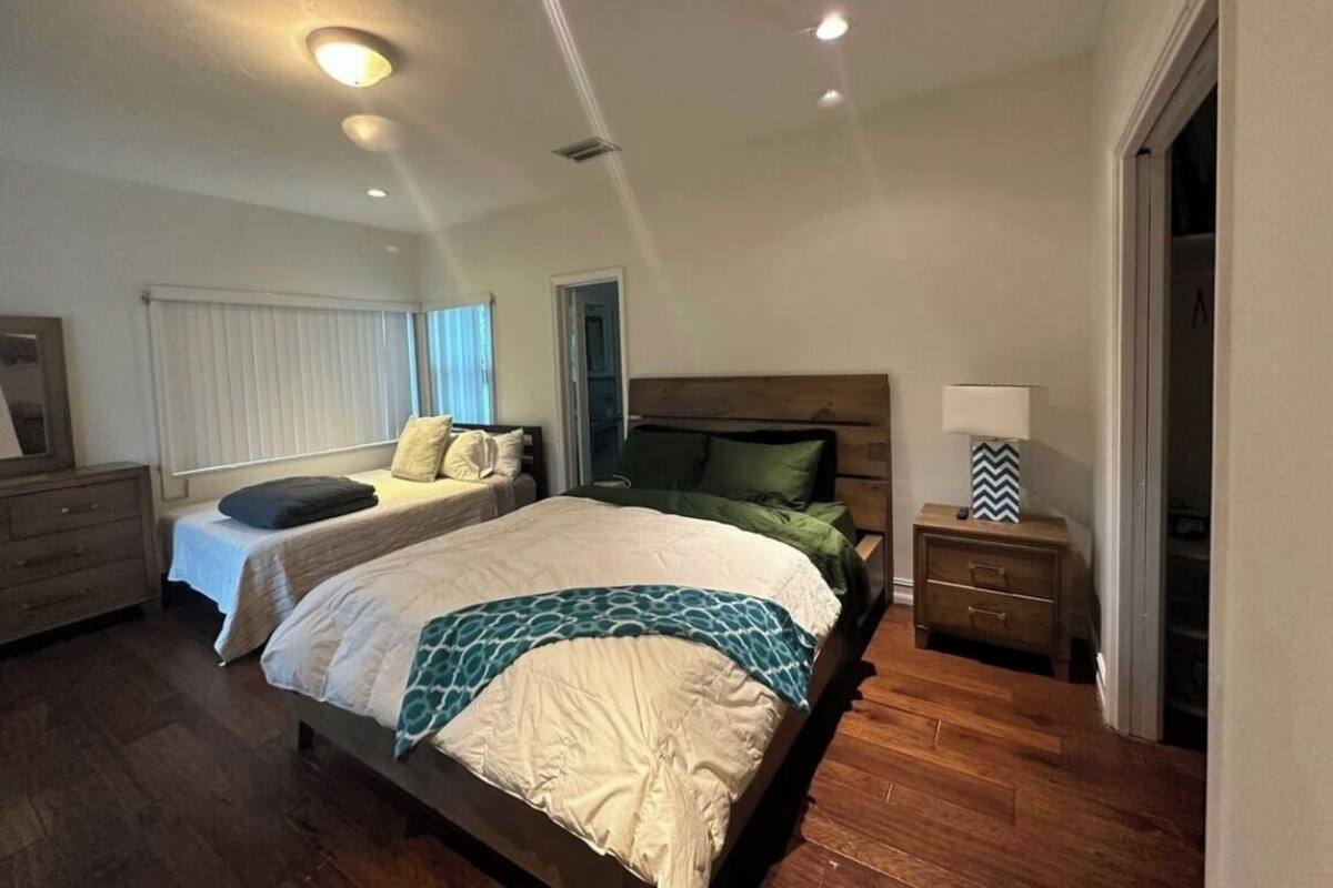 Une chambre avec deux lits, un plancher en bois, une table de chevet avec une lampe et une fenêtre avec des stores.