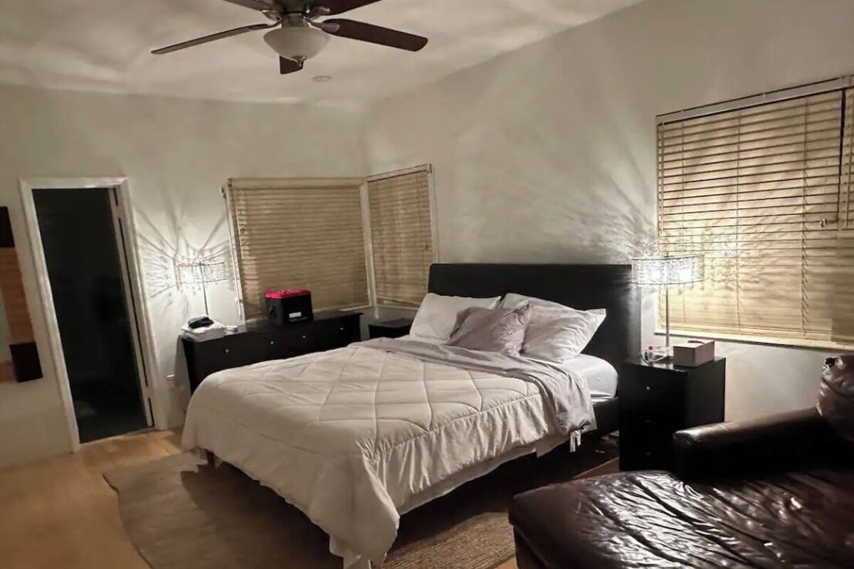 Une chambre à coucher comprenant un lit, un ventilateur de plafond, une table de chevet, une commode, une fenêtre avec des stores et un canapé en cuir.