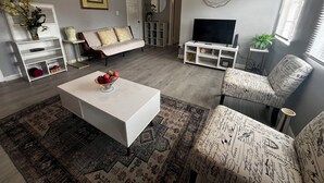 A living room with a white sofa, a coffee table, a patterned armchair, a TV stand, and a decorative wall clock.