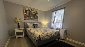 A bedroom with a bed, nightstand, lamp, air conditioner, and a painting on the wall.