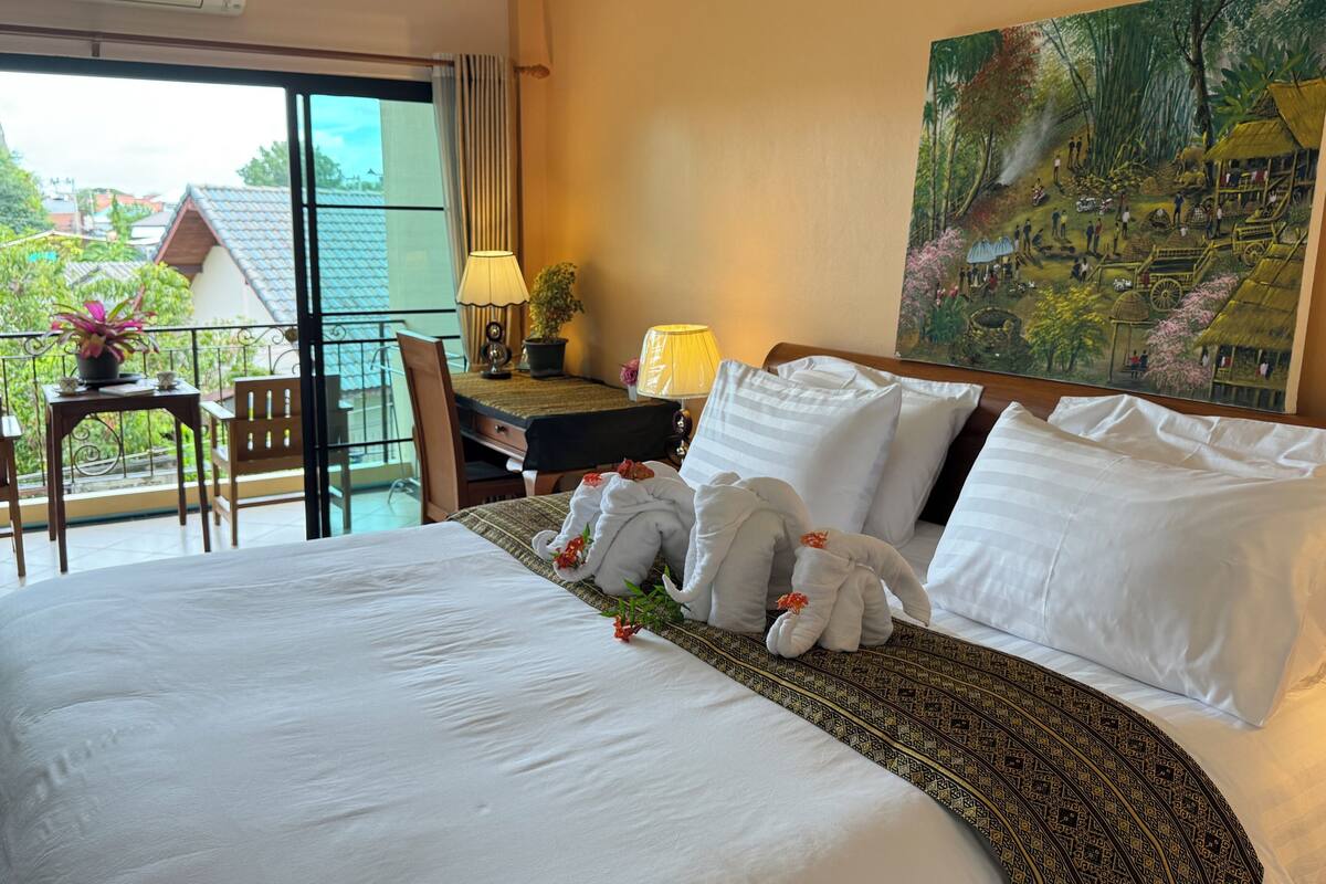 A hotel room with a large bed, a desk, a chair, a balcony with a view of buildings and greenery, and a painting on the wall.