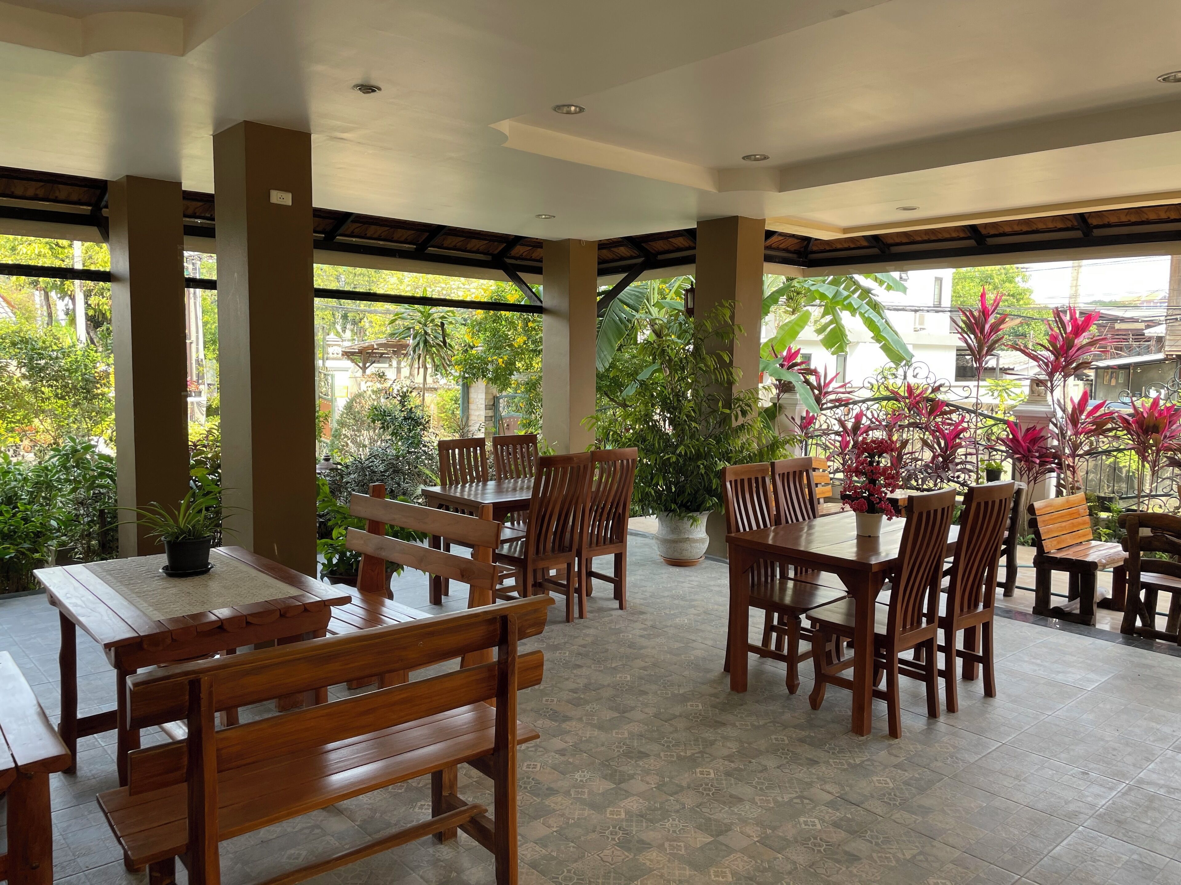 A covered outdoor seating area with wooden tables and benches, surrounded by potted plants and a view of a residential area.