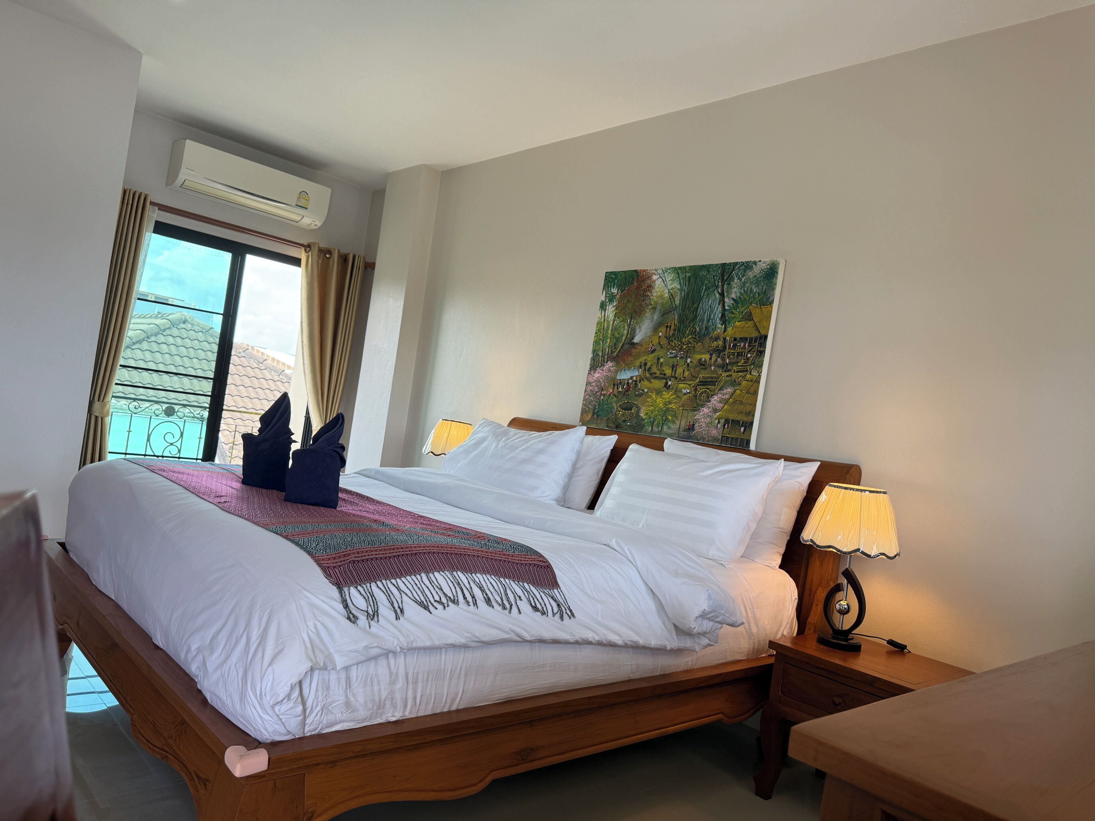 A hotel room with a large bed, a painting on the wall, an air conditioning unit, and a window with curtains.