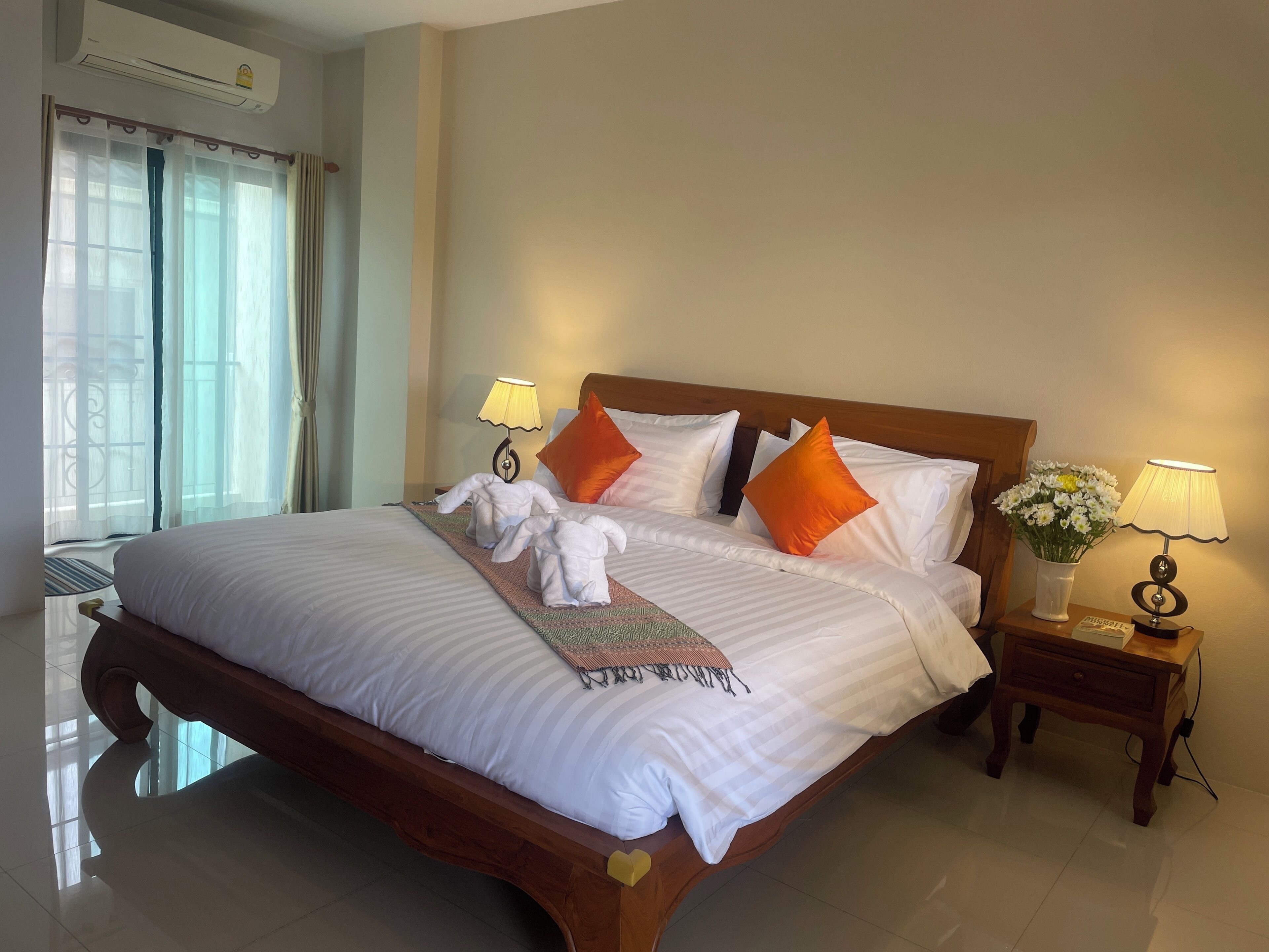 Una cama bien hecha con ropa blanca, dos almohadas naranjas y un par de toallas blancas dobladas al pie. Hay dos lámparas de noche en mesitas de madera, una con pantalla blanca y la otra con pantalla amarilla. En la mesita a la derecha hay un pequeño florero con flores blancas.
