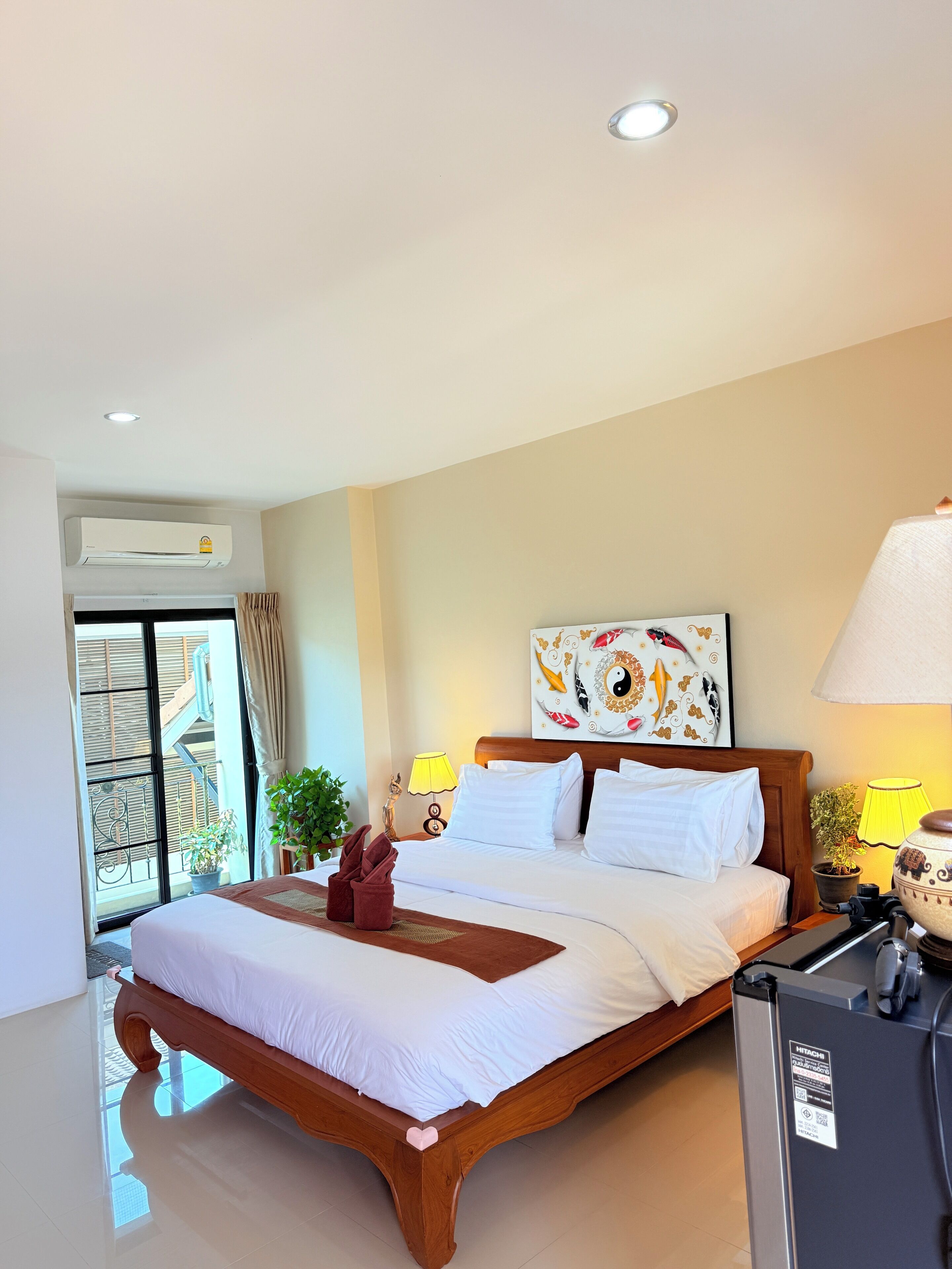 Habitación de hotel con una cama grande, ventilador de techo, aire acondicionado instalado en la pared, ventana con cortinas, una planta en maceta, un cuadro y una mesita de noche con lámpara.