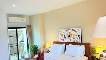Habitación de hotel con una cama grande, ventilador de techo, aire acondicionado instalado en la pared, ventana con cortinas, una planta en maceta, un cuadro y una mesita de noche con lámpara.