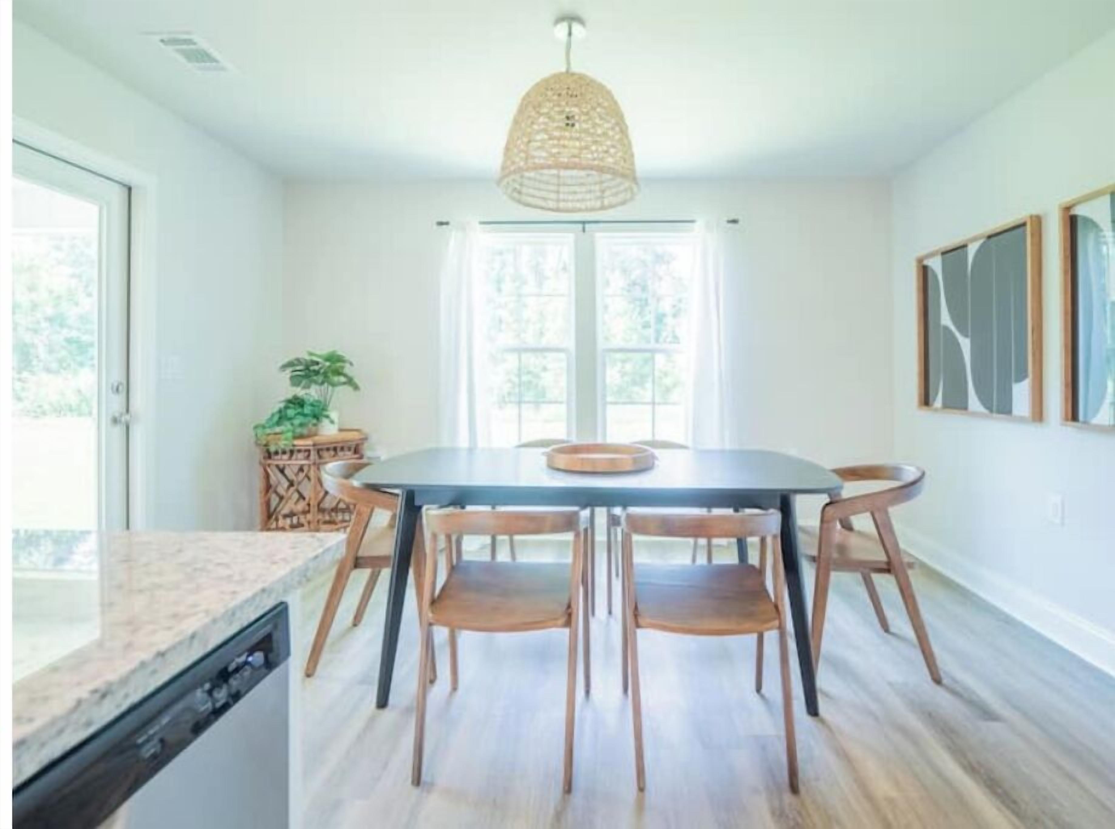 Zona pranzo con tavolo rotondo, sei sedie di legno, lampada a sospensione intrecciata e vista sul verde esterno.