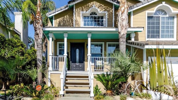 A two-story house with a front porch, a dark front door, and a small front yard with palm trees and shrubs.