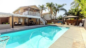 A swimming pool with a covered area, a two-story house, and palm trees in the background.