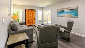 A living room with a grey sofa, a coffee table, and two armchairs. There is a wooden door and a painting on the wall.