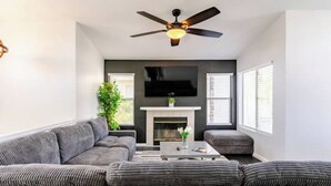 A modern living room with a grey sofa set, a fireplace, a flat-screen TV, and a ceiling fan.