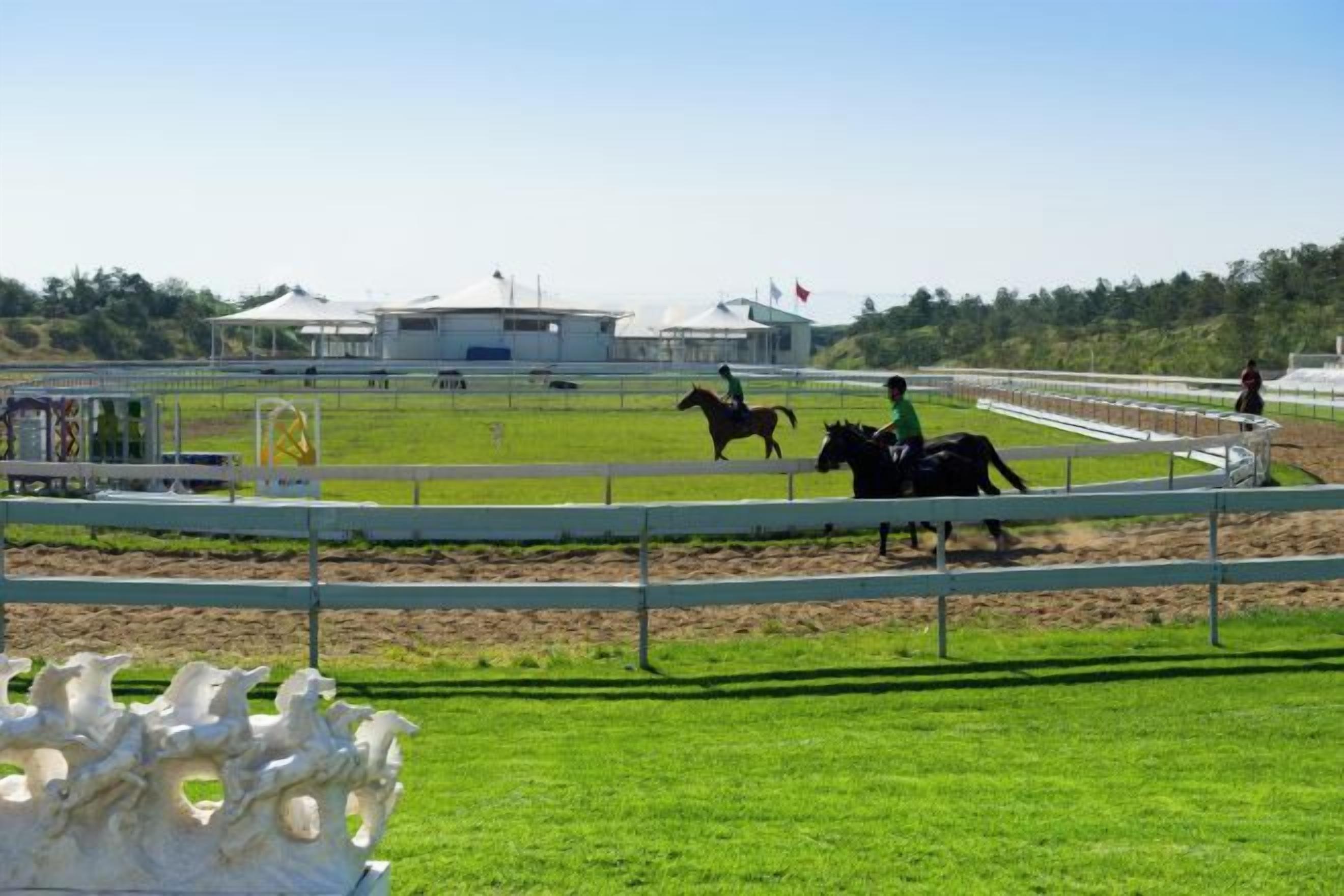 A horse racing track with a white fence, a jockey on a horse, and a stable building in the background.