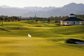 A golf course with a flagstick, a building with a dark roof, and mountains in the background. - Reignwood Pine Valley (Beijing)