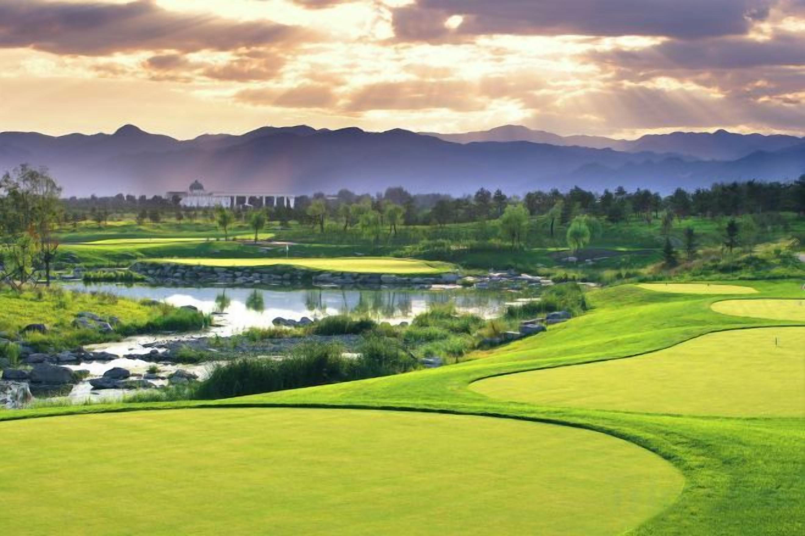 Un parcours de golf avec un étang, des arbres et des montagnes en arrière-plan.