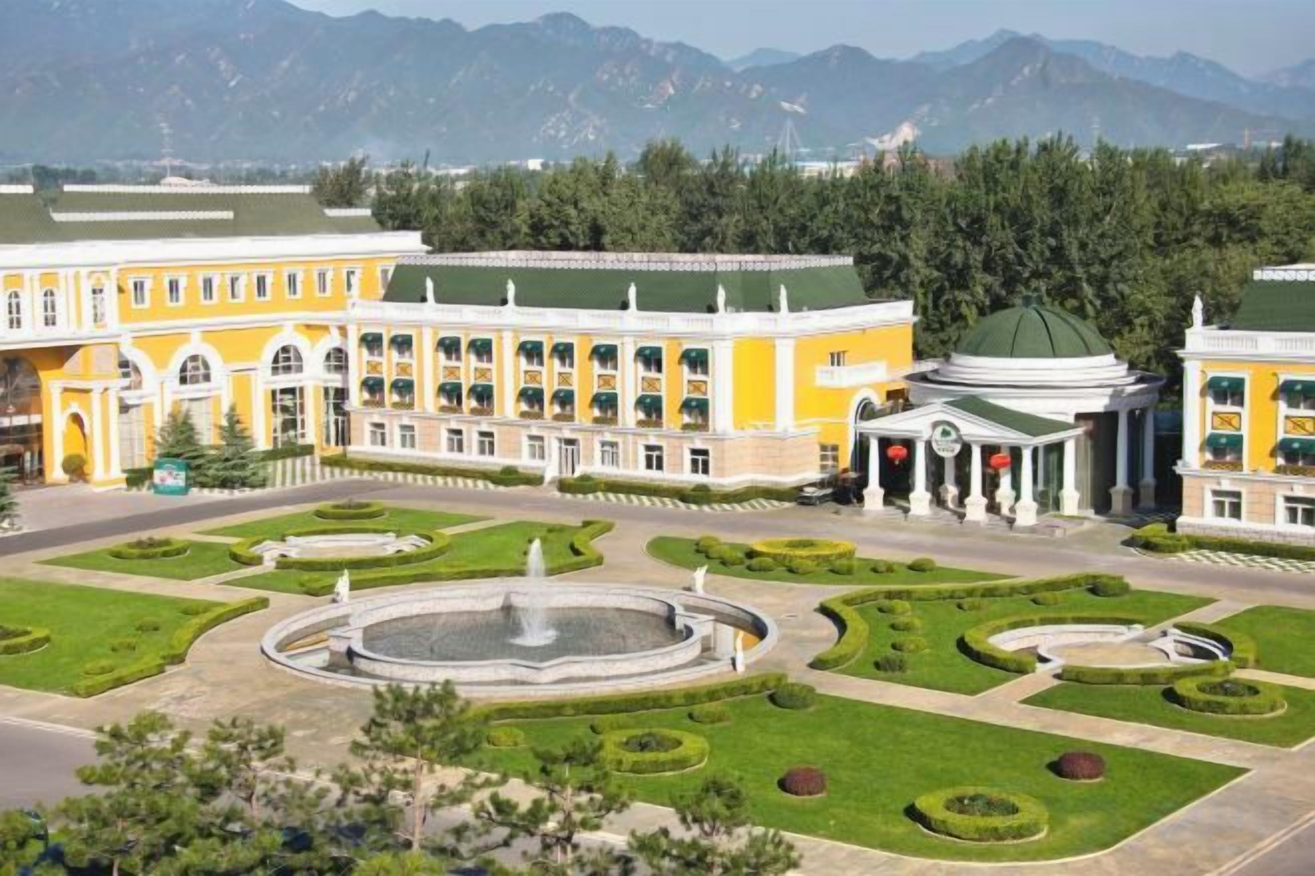 A large building with a central fountain, surrounded by manicured gardens and a mountainous backdrop.