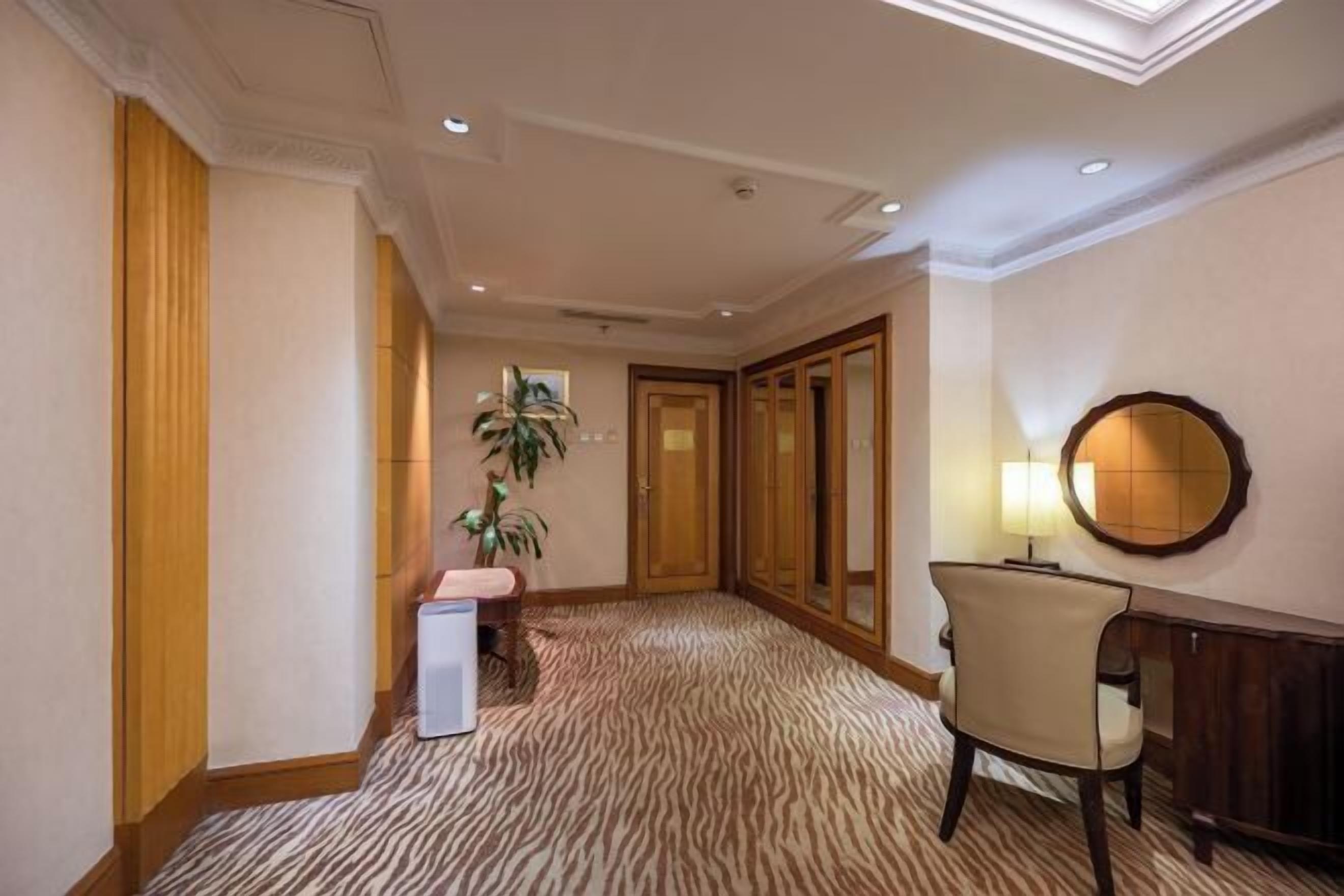 Een hotelgang met een kamerbreed tapijt, houten deuren, een bureau met lamp en een potplant.