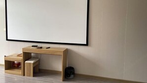A room with a large blank TV screen mounted on the wall, a wooden desk with a chair, and a small shelf with a red object. - Yanyangtian Business Hotel (Yongzhou)