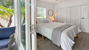 Un dormitorio con cama, mesita de noche, lámpara, armario y vistas a palmeras a través de un ventanal.