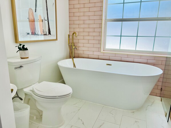 Un baño con inodoro blanco, una bañera independiente, una ventana y suelo de baldosas.