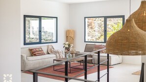A modern living room with a sofa, a coffee table, and two large windows offering a view of the outdoors. - Villa IKAL by Abahana Villas (Moraira)