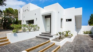 Um edifício moderno e branco com teto plano, entrada central e calçada pavimentada que leva a um conjunto de degraus.