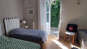 A bedroom with two beds, a radiator, a small table with a lamp, a suitcase, and a view of the outside. - House with garden near the beach of Arcachon (Arcachon)