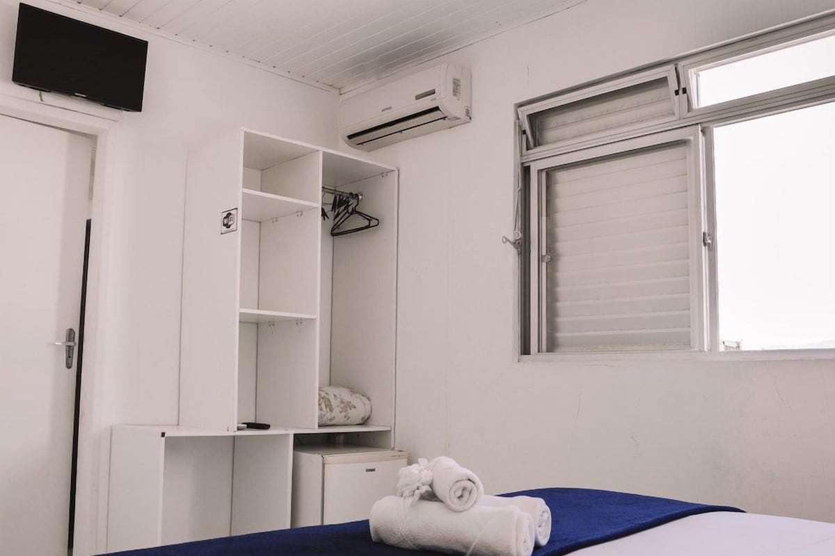 A bedroom with a bed, a wardrobe, a desk, an air conditioner, and a window with blinds.