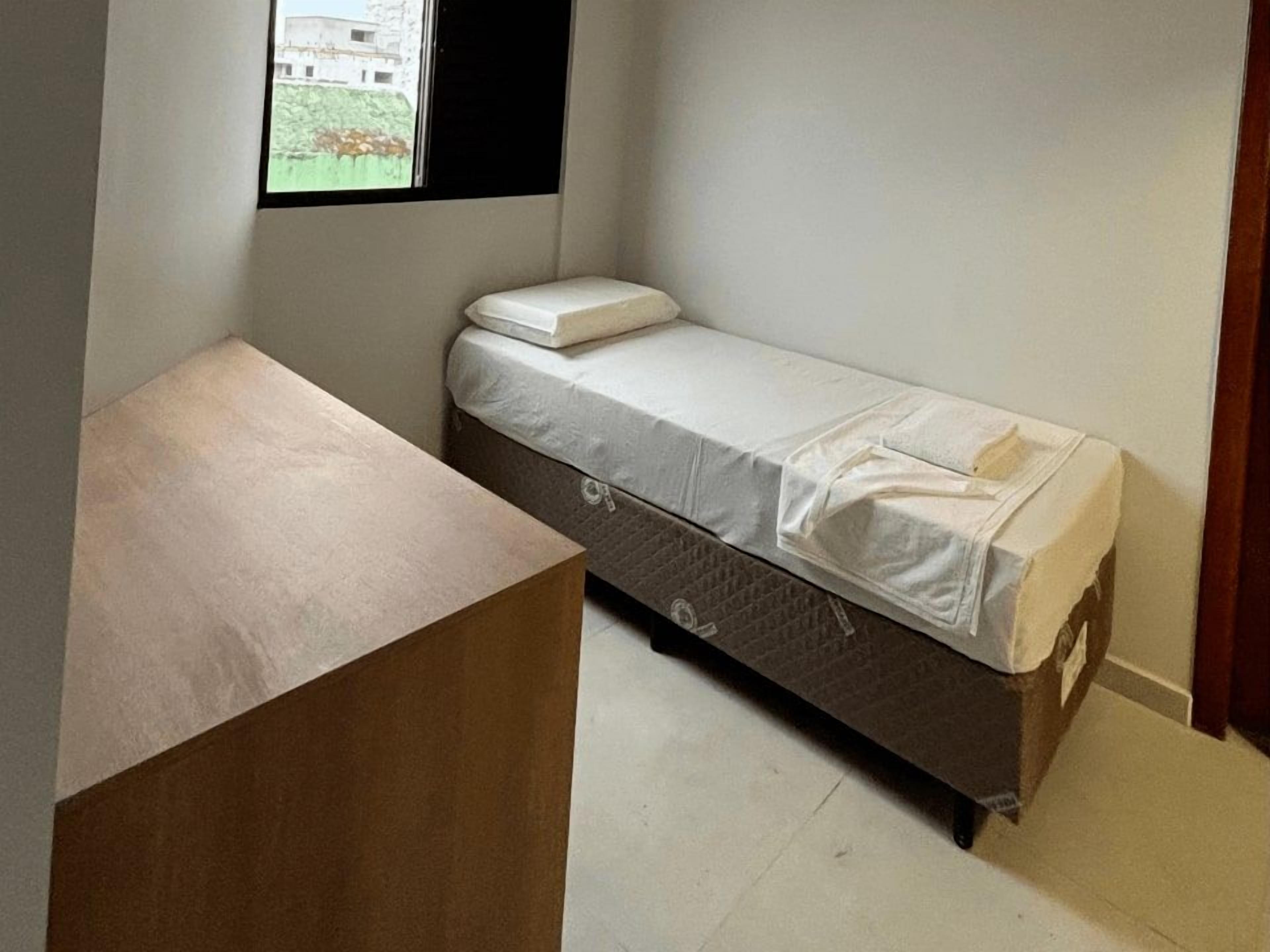 A small, single-bed room with a wooden desk and a window overlooking a building.