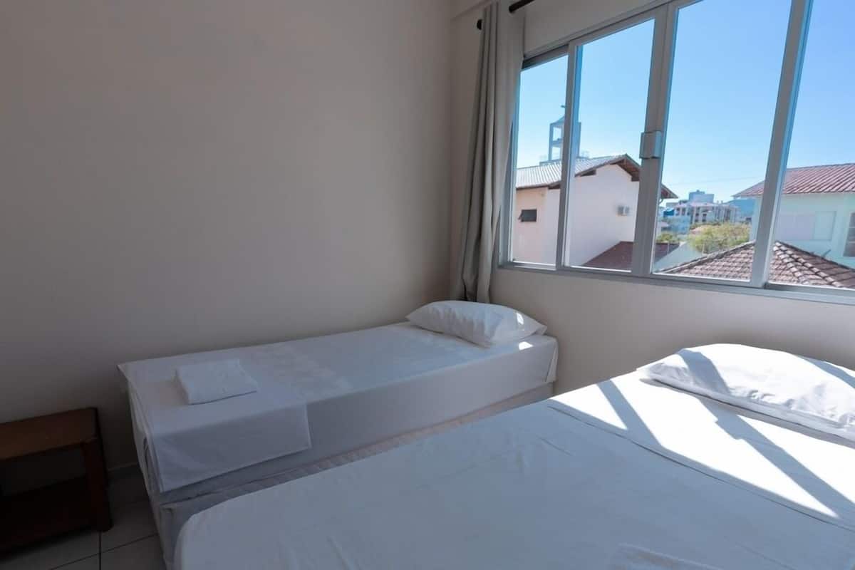 A room with two beds, a window with a view of buildings, and a bench.
