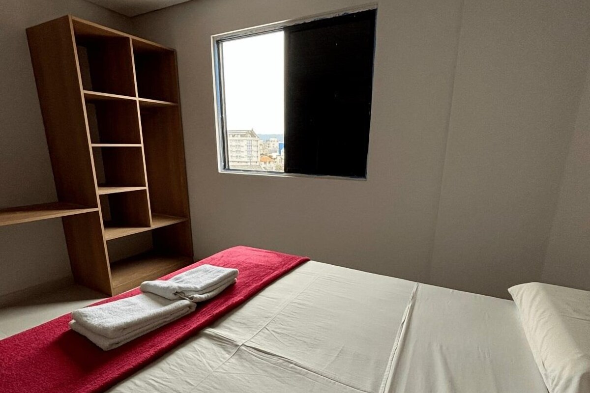 A bedroom with a bed, a red towel, a wooden wardrobe, and a window with a view of buildings.
