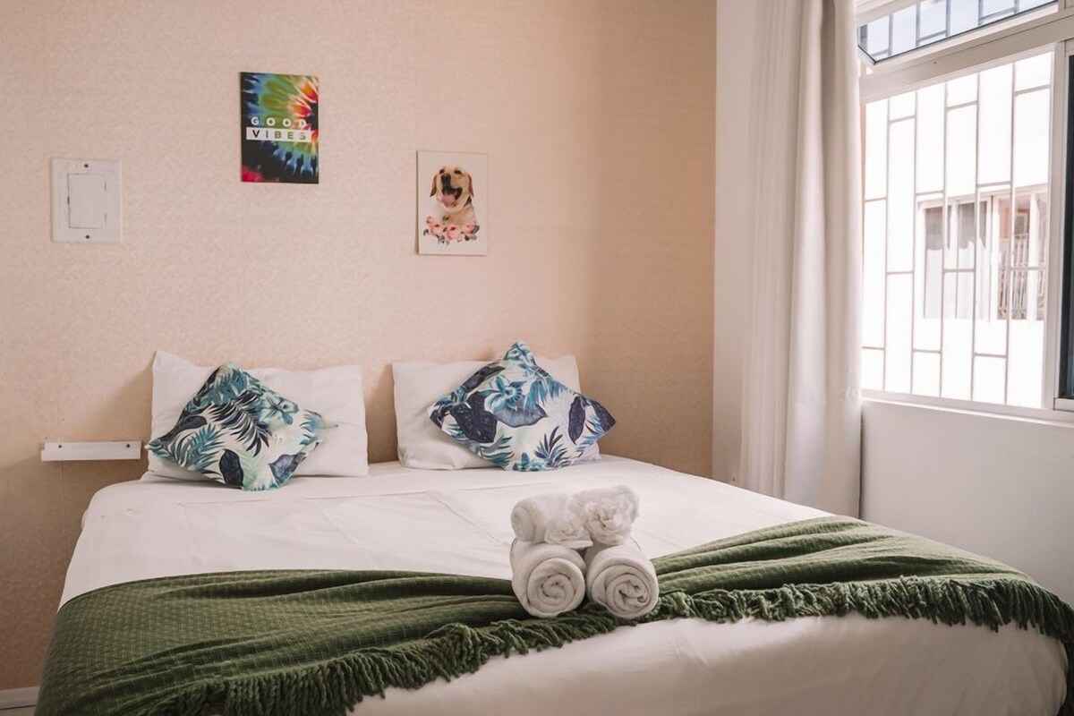 A neatly made bed with a green throw blanket and rolled towels, two tropical-themed pillows, a wall art with a dog, and a window with white curtains.
