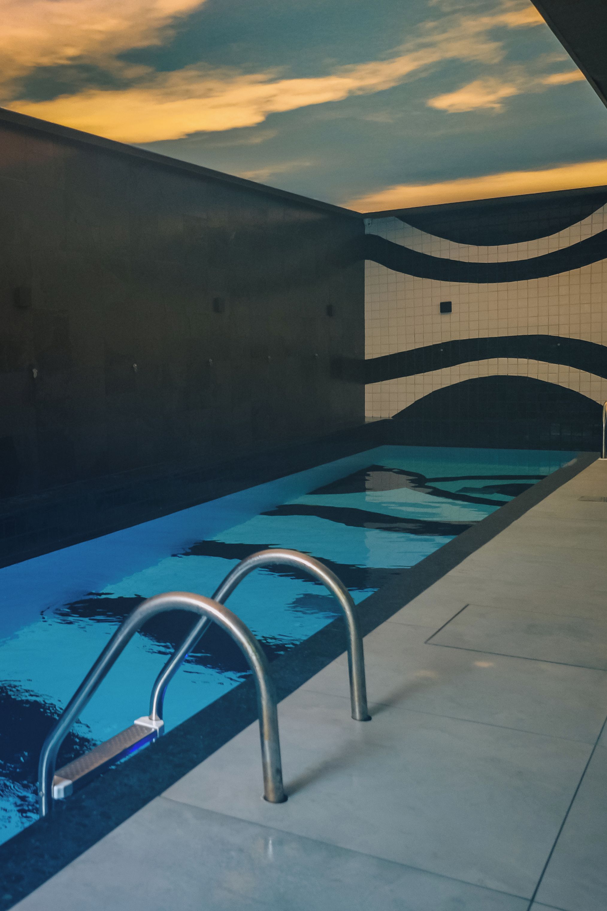 An indoor swimming pool with clear water, metal handrails, and tiled walls.