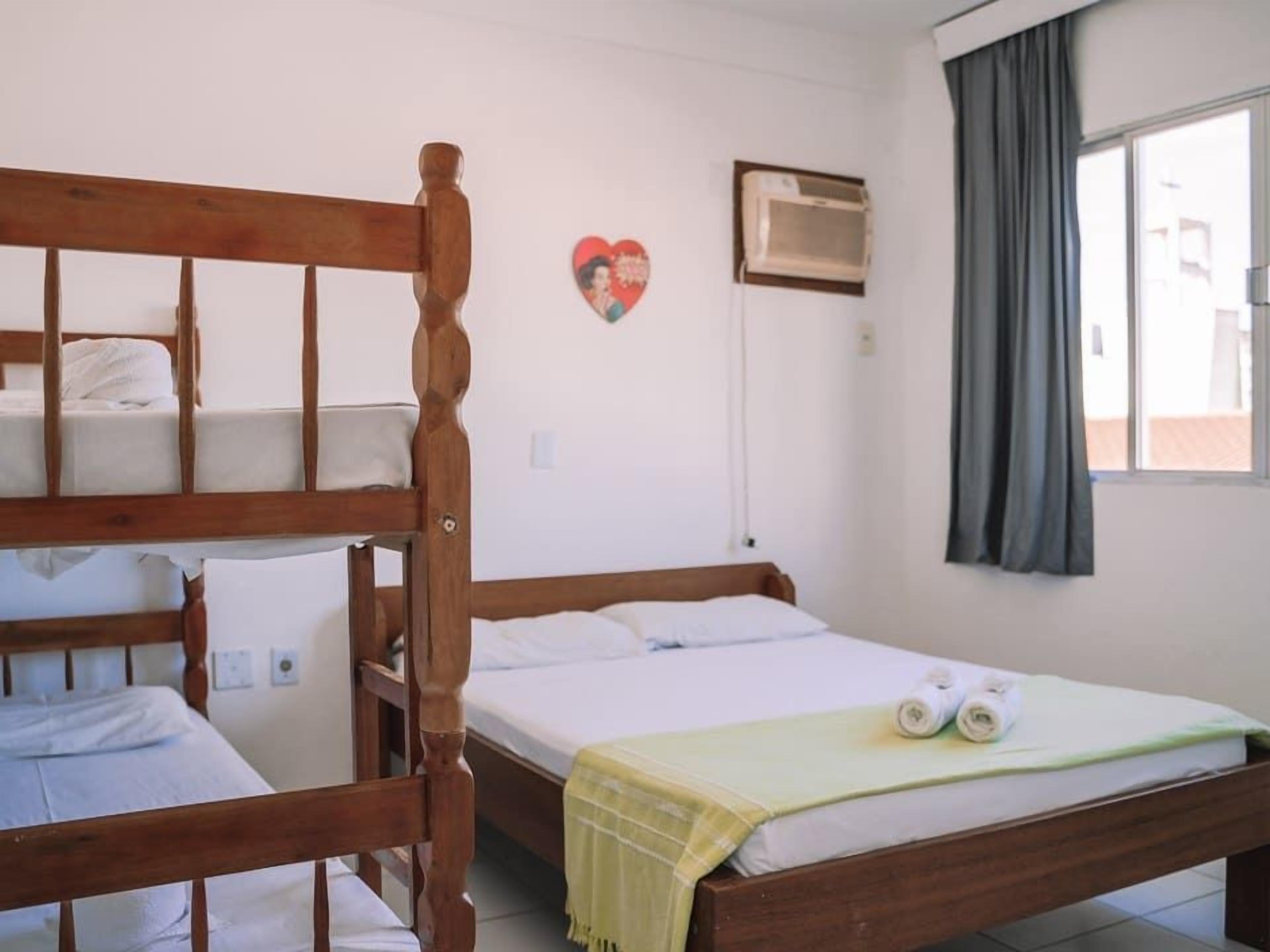 Une chambre avec un lit superposé, un lit simple, un climatiseur fixé au mur, une fenêtre avec des rideaux et une décoration en forme de cœur.