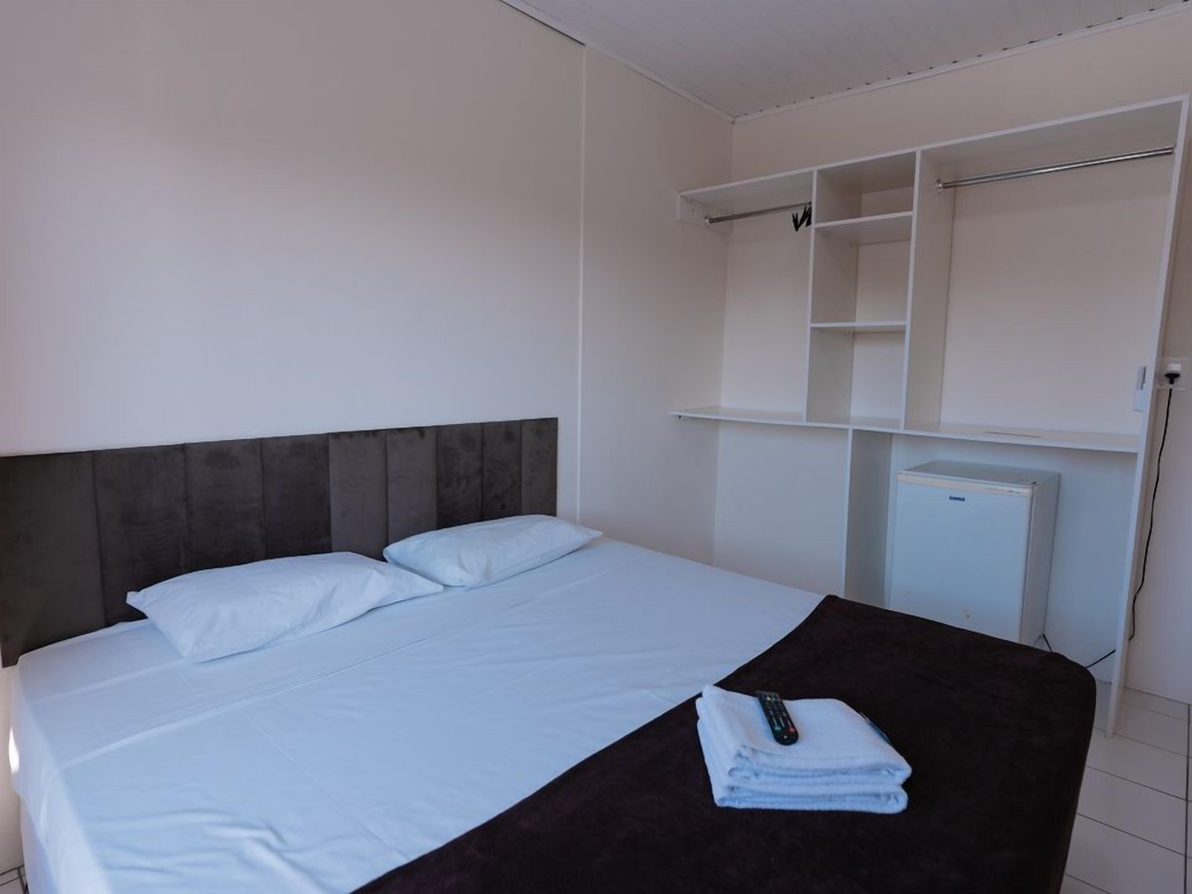 Habitación de hotel con cama, cabecera oscura, mesita de noche blanca con control remoto y una toalla doblada, y armario blanco con estantes abiertos.