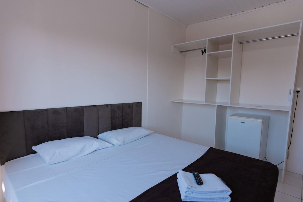 A hotel room with a bed, a dark headboard, a white bedside table with a remote control and a folded towel, and a white wardrobe with open shelves.