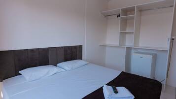 Habitación de hotel con cama, cabecera oscura, mesita de noche blanca con control remoto y una toalla doblada, y armario blanco con estantes abiertos.