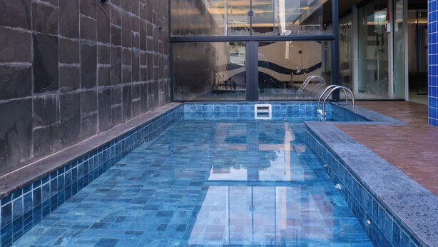 Een klein binnenzwembad met blauwe tegels, een metalen trap en een glazen wand op de achtergrond.