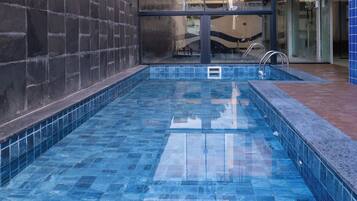 Una pequeña piscina interior con azulejos azules, una escalera de metal y una pared de vidrio al fondo.