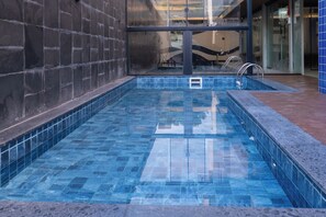 Una pequeña piscina interior con azulejos azules, una escalera de metal y una pared de vidrio al fondo.
